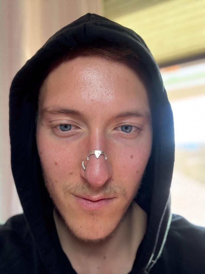 May include: A silver-colored nose ring with a geometric design. The nose ring has a triangular center piece and curved side elements. The person is wearing a black hooded sweatshirt.