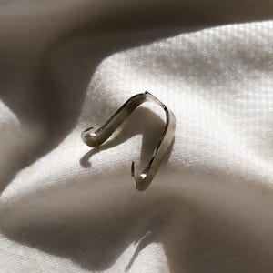 May include: A small, silver-colored metal clip with a curved design. The clip has a hook on each end and is resting on a white, textured fabric. The lighting creates shadows that highlight the clip's shape.