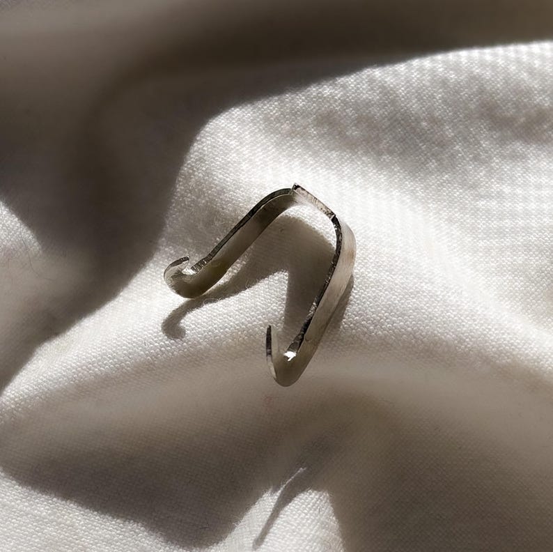 May include: A small, silver-colored metal clip with a curved design. The clip has a hook on each end and is resting on a white, textured fabric. The lighting creates shadows that highlight the clip's shape.