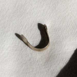 May include: A small, curved, silver-colored metal object, possibly a jewelry component or finding, rests on a white, textured surface. The object has a U-shape with a slightly flattened end and a shadow.