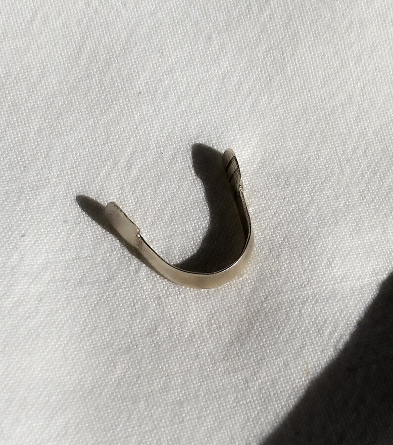 May include: A small, curved, silver-colored metal object, possibly a jewelry component or finding, rests on a white, textured surface. The object has a U-shape with a slightly flattened end and a shadow.