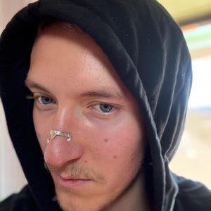 May include: A person wearing a black hooded sweatshirt with a silver nose ring. The nose ring has a geometric design. The person has fair skin, blue eyes, and a light mustache.
