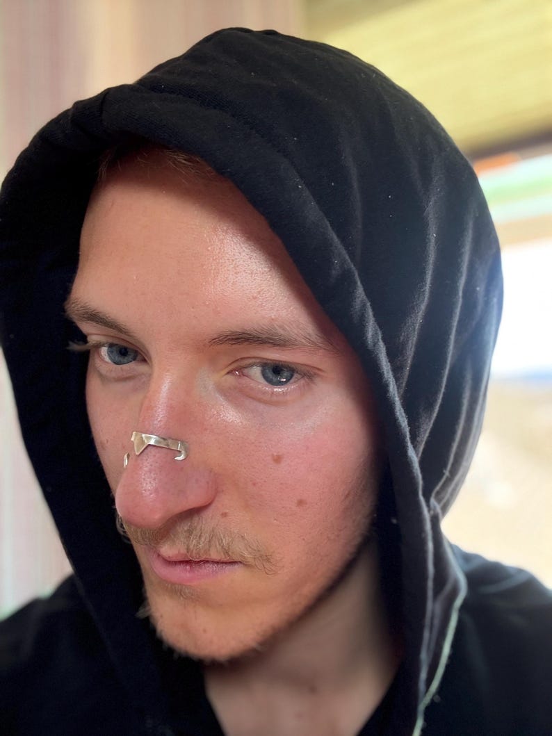 May include: A person wearing a black hooded sweatshirt with a silver nose ring. The nose ring has a geometric design. The person has fair skin, blue eyes, and a light mustache.
