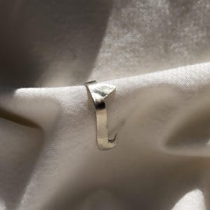 May include: A silver ring with a unique, angular design. The ring features a flat, rectangular top and a curved, hook-like end. The ring is set against a white, textured background.