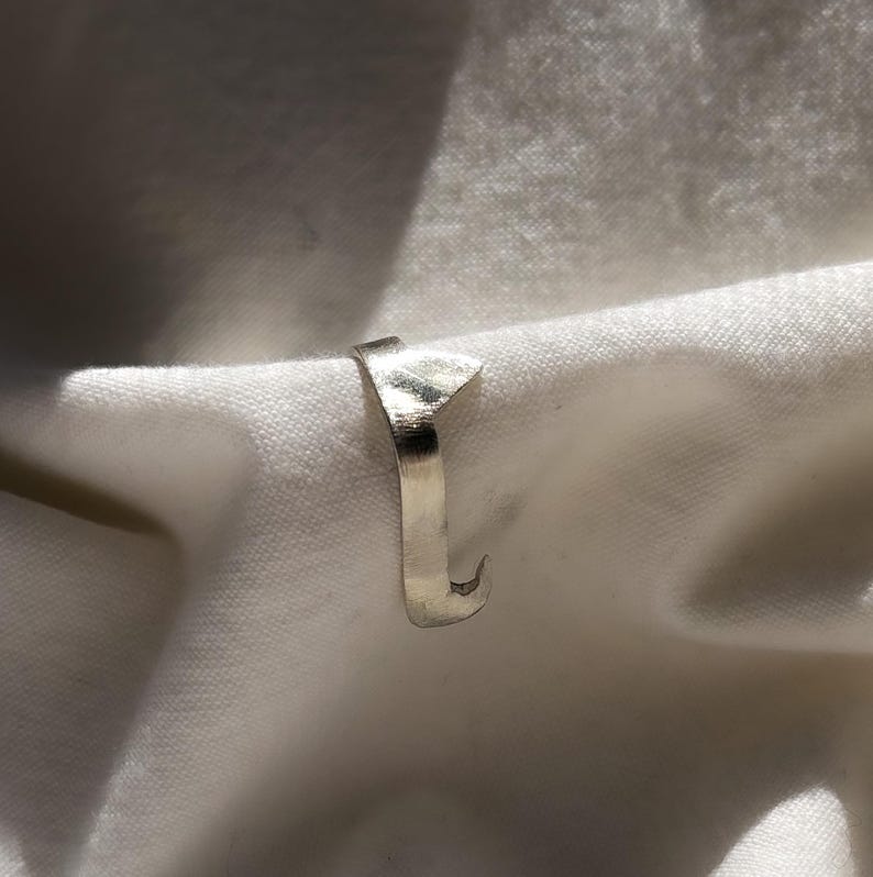May include: A silver ring with a unique, angular design. The ring features a flat, rectangular top and a curved, hook-like end. The ring is set against a white, textured background.