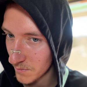 May include: A silver-colored nose ring is worn on the nose of a person wearing a black hooded sweatshirt. The nose ring has a unique design. The person has fair skin, blue eyes, and light brown hair.
