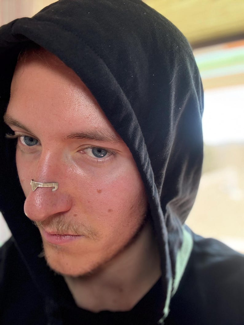 May include: A silver-colored nose ring is worn on the nose of a person wearing a black hooded sweatshirt. The nose ring has a unique design. The person has fair skin, blue eyes, and light brown hair.