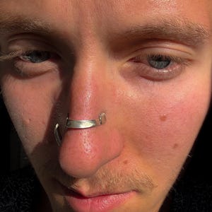 May include: Close-up of a person's face with a silver nose ring. The nose ring is a curved bar that sits across the septum. The skin is fair with visible pores and some freckles. The eyes are blue and the lighting is warm.