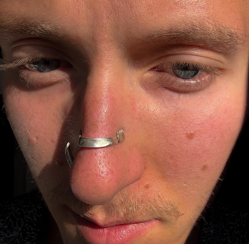 May include: Close-up of a person's face with a silver nose ring. The nose ring is a curved bar that sits across the septum. The skin is fair with visible pores and some freckles. The eyes are blue and the lighting is warm.