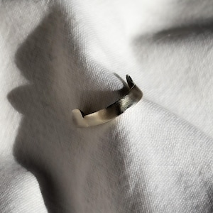 May include: A silver-toned ring with a unique, curved design. The ring is set against a white, textured fabric, with shadows adding depth. The ring's surface has a textured appearance, reflecting light. This piece of jewelry is a minimalist accessory.