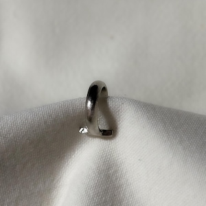 May include: A silver-colored ring with a curved top and a flat, rectangular bottom. The ring is set on a white fabric background. The ring has a small, clear gemstone on the bottom.