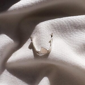 May include: A silver-colored ring with a unique, architectural design. The ring features a geometric shape with a pointed top and a curved band. The ring is set against a white, textured background, creating a contrast that highlights its metallic sheen.