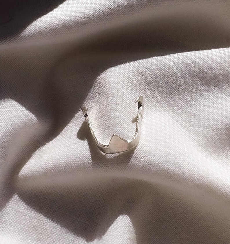 May include: A silver-colored ring with a unique, architectural design. The ring features a geometric shape with a pointed top and a curved band. The ring is set against a white, textured background, creating a contrast that highlights its metallic sheen.