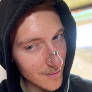 May include: A silver nose ring with a geometric design. The nose ring is worn on the nose of a person wearing a black hooded sweatshirt. The person has fair skin, blue eyes, and reddish-brown hair.