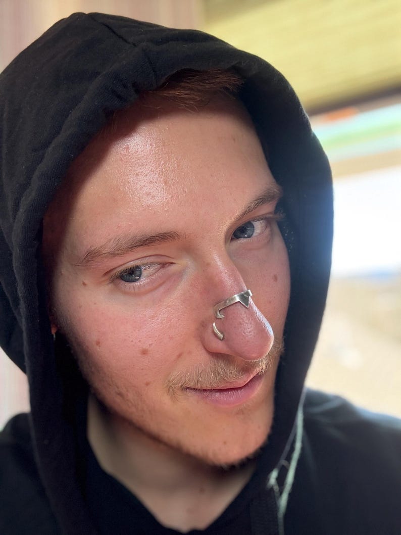 May include: A silver nose ring with a geometric design. The nose ring is worn on the nose of a person wearing a black hooded sweatshirt. The person has fair skin, blue eyes, and reddish-brown hair.