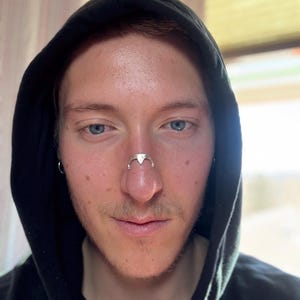 May include: A silver-colored nose ring with a triangular design is worn on the nose. The person is wearing a black hooded sweatshirt. The background is blurred, with natural light coming from a window.