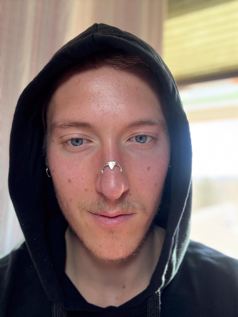 May include: A silver-colored nose ring with a triangular design is worn on the nose. The person is wearing a black hooded sweatshirt. The background is blurred, with natural light coming from a window.
