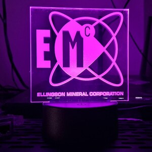 May include: A square, illuminated sign with the text "EMC" in large, bold letters, surrounded by a circular design. The words "Ellingson Mineral Corporation" are printed below. The sign is lit with a purple glow and sits on a black base.