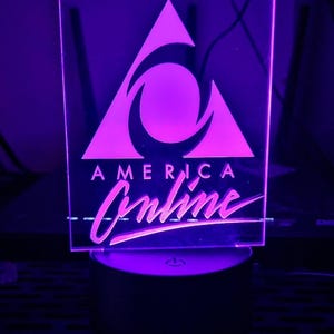 May include: A square, illuminated sign with the America Online logo. The sign is made of clear acrylic and glows with a purple light. The logo features a triangle and a stylized swirl, with the words "AMERICA Online" below.