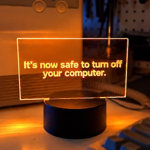 May include: A rectangular acrylic light panel with a black circular base, emitting a warm orange glow. The panel features the text 'It's now safe to turn off your computer.' in a clear, sans-serif font, illuminated against the dark room.