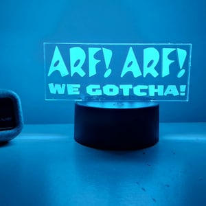 May include: A glowing LED sign with the text "ARF! ARF! WE GOTCHA!" in a playful font. The sign sits on a black base, next to a digital alarm clock displaying 3:39 in red numerals. The scene is bathed in blue light.