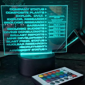 May include: A teal blue acrylic sign with a white background and black text. The sign has a list of company departments, including "Company Status", "Composite Plants", "Explor. DVLT", "Explor. Research", "Geologic Research", "Garbage", "Geologic Budgets", "Diving Consultants", "Ballast Reports", "Mine Development", "Blast Frnc. Status", "Nuclear Research", "Recruitment", and "Airfreight Status". The sign is lit up with a teal blue light and is sitting on a black base. A remote control with multiple colored buttons is in the foreground.