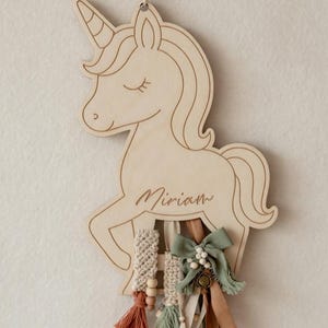 Könnte beinhalten: Wanddekoration aus Holz in Form eines Einhorns mit dem eingravierten Namen "Miriam". Das Einhorn hat eine fließende Mähne und ein Horn. Es ist mit Makramee, Quasten, Bändern und einer Schleife in Beige-, Grün- und Brauntönen verziert.