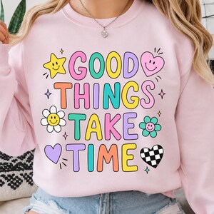 Może przedstawiać: Jasnoróżowa bluza z okrągłym dekoltem i kolorowym napisem GOOD THINGS TAKE TIME. Wzór zawiera żółtą gwiazdkę, różowe serce, biały kwiatek, turkusowy kwiatek, fioletowe serce oraz czarno-białe serce w szachownicę, wszystkie z uśmiechniętymi buźkami.