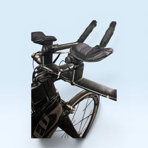 May include: Black triathlon bicycle with aero bars, padded armrests, and a sleek design. The bicycle features a black frame, wheels, and components, with a focus on aerodynamic efficiency for speed and performance. The image is set against a light blue background.