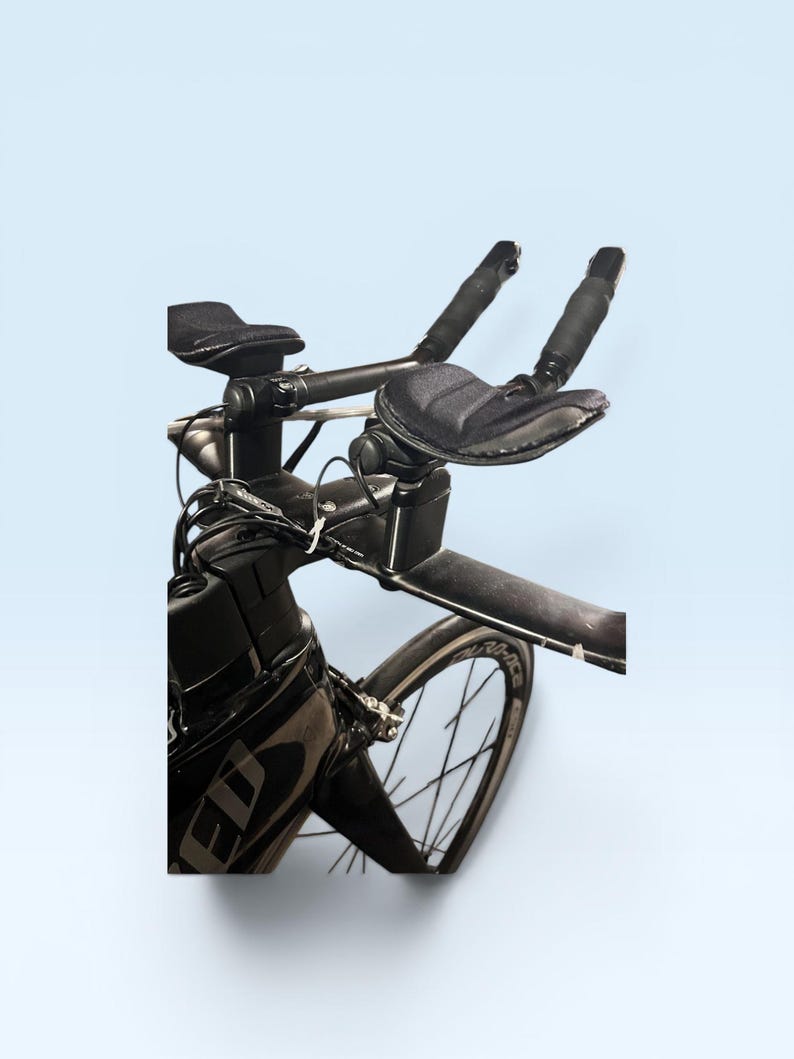 May include: Black triathlon bicycle with aero bars, padded armrests, and a sleek design. The bicycle features a black frame, wheels, and components, with a focus on aerodynamic efficiency for speed and performance. The image is set against a light blue background.
