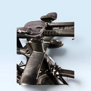 May include: Close-up of a black road bicycle handlebar with padded armrests and a sleek design. The handlebars have white measurement markings. The bike is set against a light blue background.
