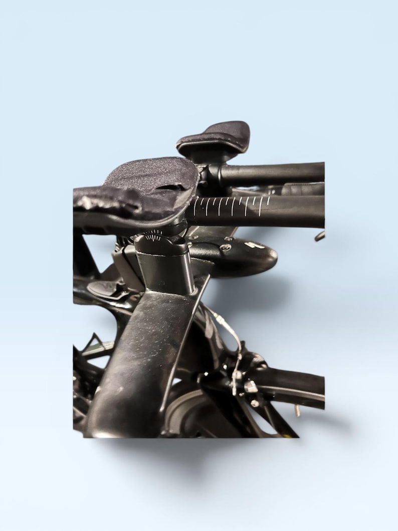 May include: Close-up of a black road bicycle handlebar with padded armrests and a sleek design. The handlebars have white measurement markings. The bike is set against a light blue background.