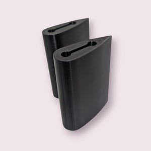 May include: Two black, aerodynamic-shaped objects with a slot cut into the top. The objects are positioned next to each other, slightly overlapping. The background is a soft, pale pink.