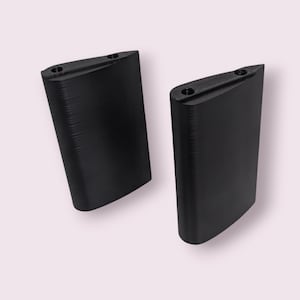 Puede incluir: Dos objetos negros en forma de ala con una superficie texturizada y dos pequeños agujeros en la parte superior. Los objetos están colocados uno al lado del otro sobre un fondo rosa claro. El diseño es elegante y moderno.