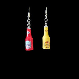 Puede incluir: Un par de pendientes originales con forma de botellas de ketchup y mostaza en miniatura. La botella de ketchup roja tiene texto blanco y una etiqueta, mientras que la botella de mostaza amarilla tiene una etiqueta amarilla. Ambos cuelgan de ganchos plateados.