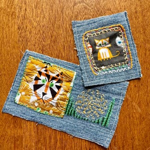 May include: Three denim patches with embroidered designs. One patch features a tiger face, another a cat, and the third has an abstract design with green grass. The patches are square and have a stitched border.