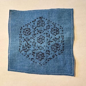 May include: A blue denim square with a black embroidered design of a geometric pattern with repeating flower shapes.