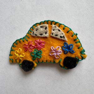 May include: A handmade felt car in a bright yellow color, adorned with colorful embroidered flowers. The car features black wheels, off-white windows, and green stitching around the edges. This whimsical design is perfect for children's crafts or as a decorative element.