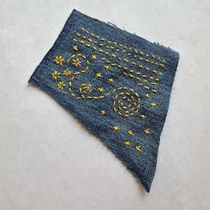 May include: A piece of denim fabric with yellow embroidered celestial designs. The embroidery includes stars, floral shapes, and a spiral pattern. The denim is a dark blue color.