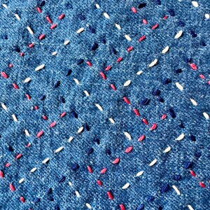Op de afbeelding: Close-up van een blauwe denim stof met witte, roze en blauwe stiksels in een diagonaal patroon.