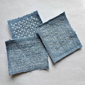 Pode incluir: Três quadrados de ganga com desenhos bordados brancos. Os padrões de bordado incluem formas de cruz e estrela. A ganga tem uma lavagem azul clara e bordas desfiadas, criando uma estética artesanal.