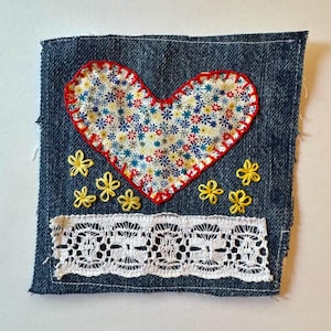 Könnte beinhalten: Ein Denim-Patch mit einem herzförmigen Stoff-Applikation mit Blumenmuster, umrandet von roten Nähten. Gelbe gestickte Blumen und eine weiße Spitzenborte verzieren den Patch und erzeugen eine handwerkliche Ästhetik. Der Patch ist quadratisch.