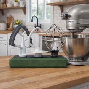 May include: A stainless steel stand mixer with a silver bowl, next to a green wooden block holding three mixer attachments: a gray paddle, a white dough hook, and a wire whisk. The kitchen setting includes wooden countertops and white cabinets.