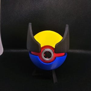 May include: A 3D-printed prop in the shape of a ball, with a yellow upper section, blue lower section, and a red band. It has a central metallic detail and two black, wing-shaped elements. The prop is displayed against a dark backdrop.