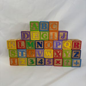 May include: A collection of wooden alphabet blocks featuring brightly coloured letters and numbers. The blocks are stacked, displaying letters A to Z and numbers 1 to 5. The letters and numbers are painted in various colours.