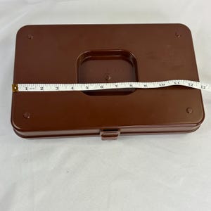 May include: A brown plastic storage box with a rectangular shape and a recessed center. The box is approximately 12 inches long. A measuring tape is placed across the top of the box.