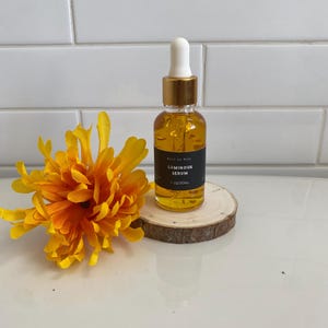 May include: A small amber glass bottle with a gold lid and a white dropper. The bottle is labeled "Luminous Serum" and contains 1 oz/30mL of product. The bottle is sitting on a small wooden round.