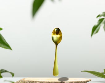 May include: A gold-colored spoon levitates above a wooden slice, surrounded by green leaves. The spoon has a rounded bowl and a slender handle. The background is a soft white, creating a minimalist aesthetic. The spoon appears to be made of metal.