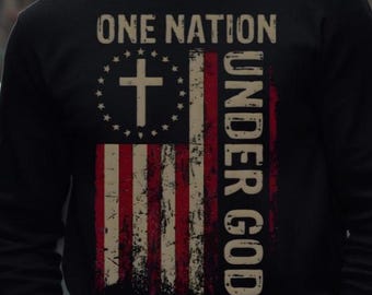 May include: Black long-sleeve shirt featuring a distressed American flag design with a cross and the text "ONE NATION UNDER GOD". The flag design incorporates red and white stripes and a blue field with a cross.