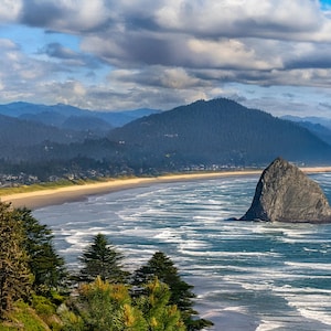 Peut inclure: Paysage côtier avec une plage de sable, des vagues océaniques et une grande formation rocheuse dans l'eau. Des montagnes et un ciel nuageux complètent la scène naturelle. Des arbres verts luxuriants sont au premier plan.